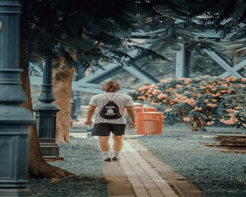 Rear view of a modern individual walking down a clean pathway in a sunlit green public park with city architecture blurred in background